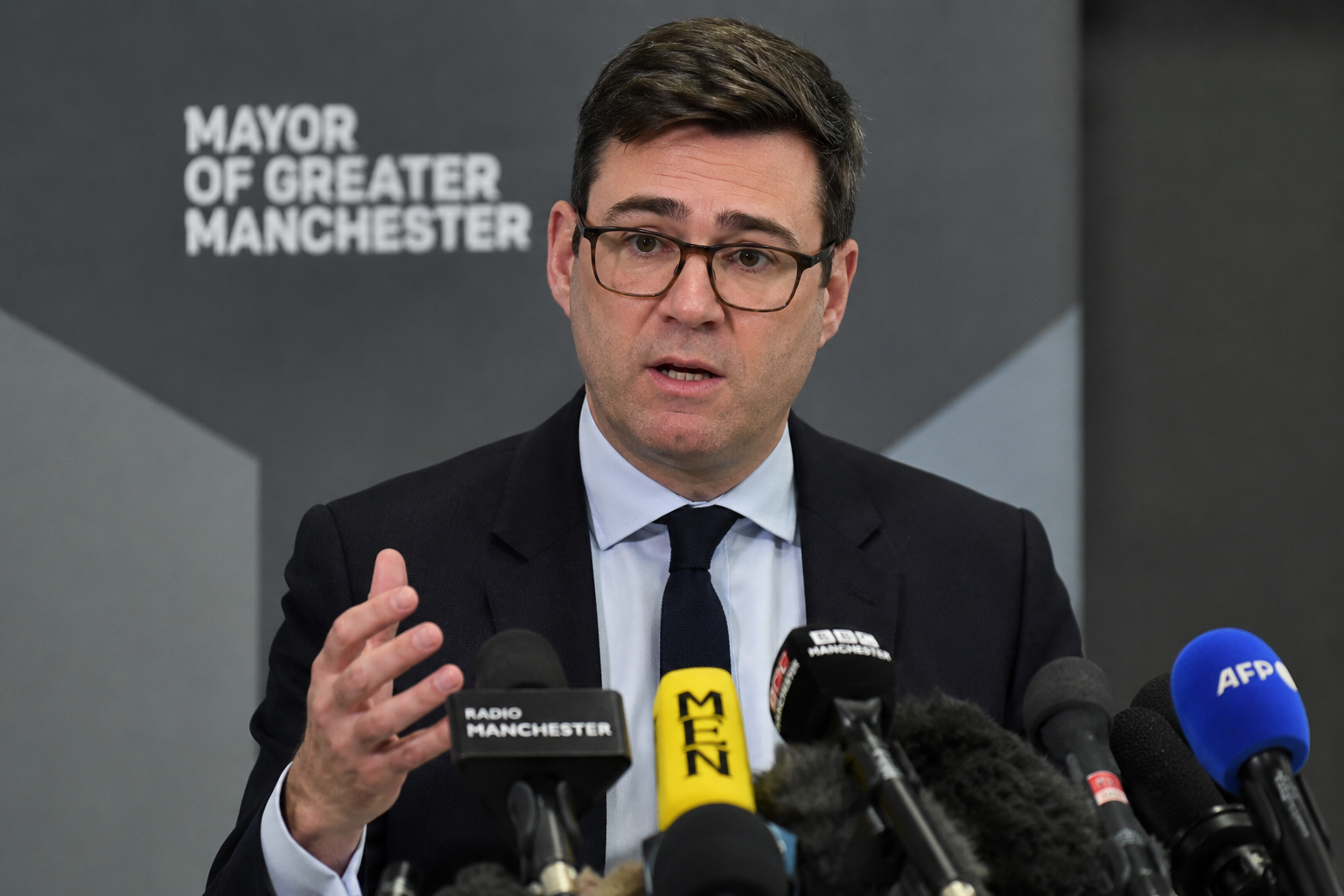 British politician Andy Burnham speaking at a press conference with media microphones in front under cinematic lighting.