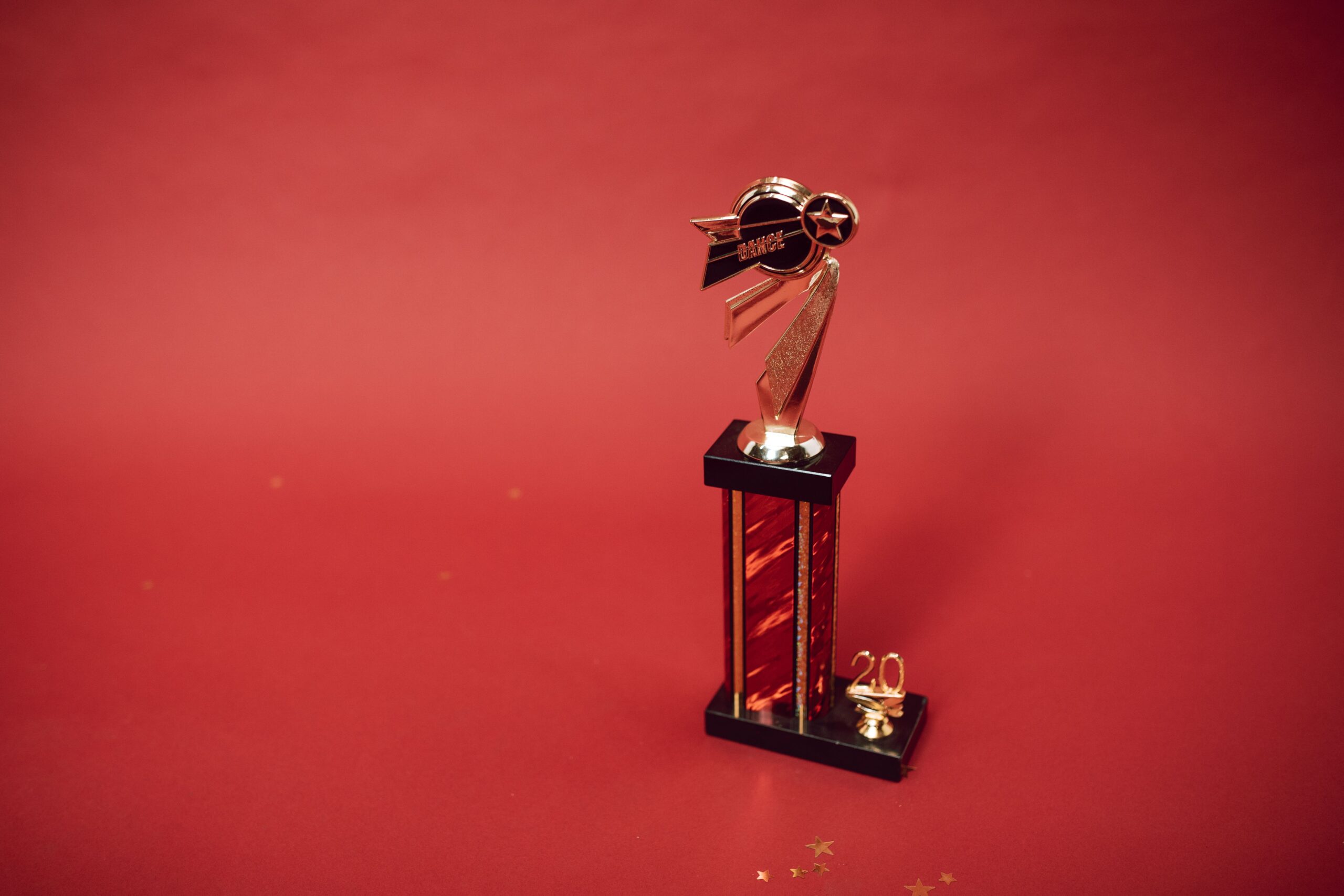 A professional entertainment journalism photograph showcasing a glittering golden award statue layout against a blurry red carpet media backdrop