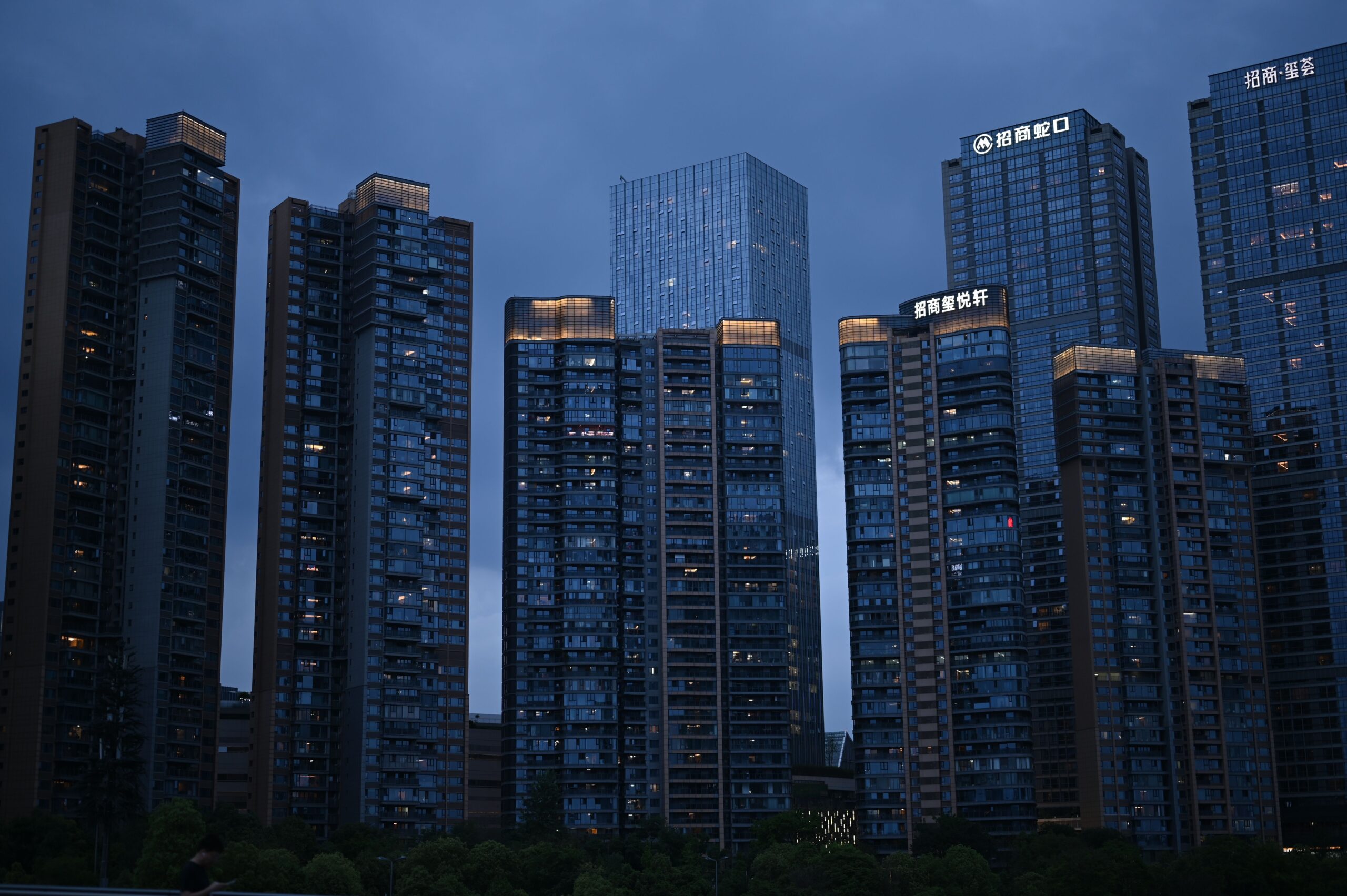 A professional financial journalism photograph showcasing architectural blueprints and commercial real estate documents laid out cleanly on a modern executive glass desk with blurred corporate skyscrapers in the background