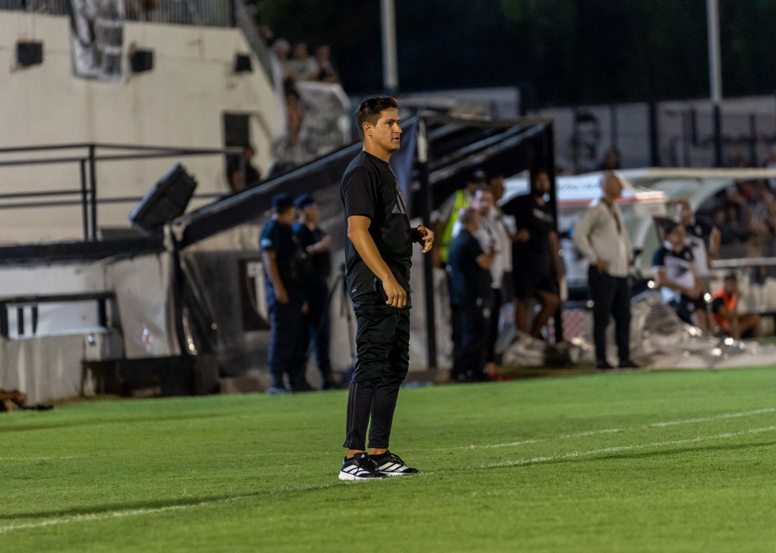 A professional football manager standing on the stadium pitch sideline under bright arena lights during a matc