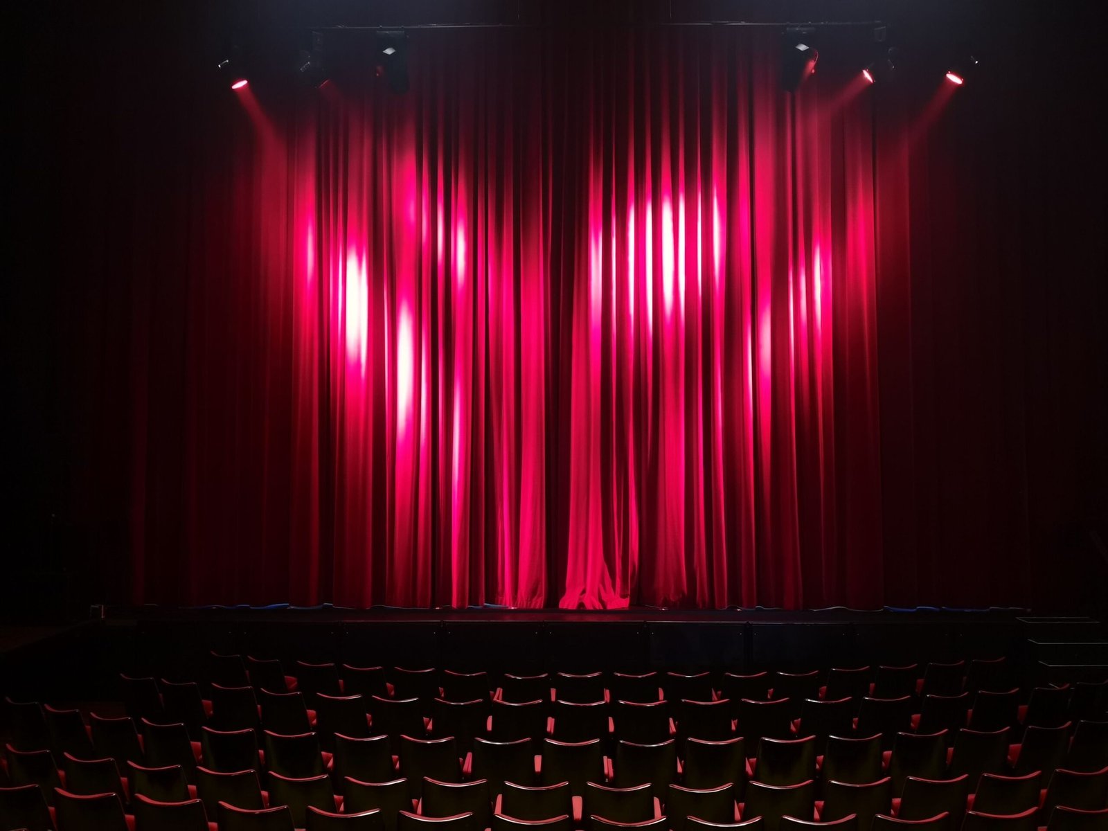 A professional entertainment journalism photograph showcasing a dramatic empty cinema hall with deep red velvet curtains and golden stage lights honoring a tribute after Sam Neill Jurassic Park actor dies
