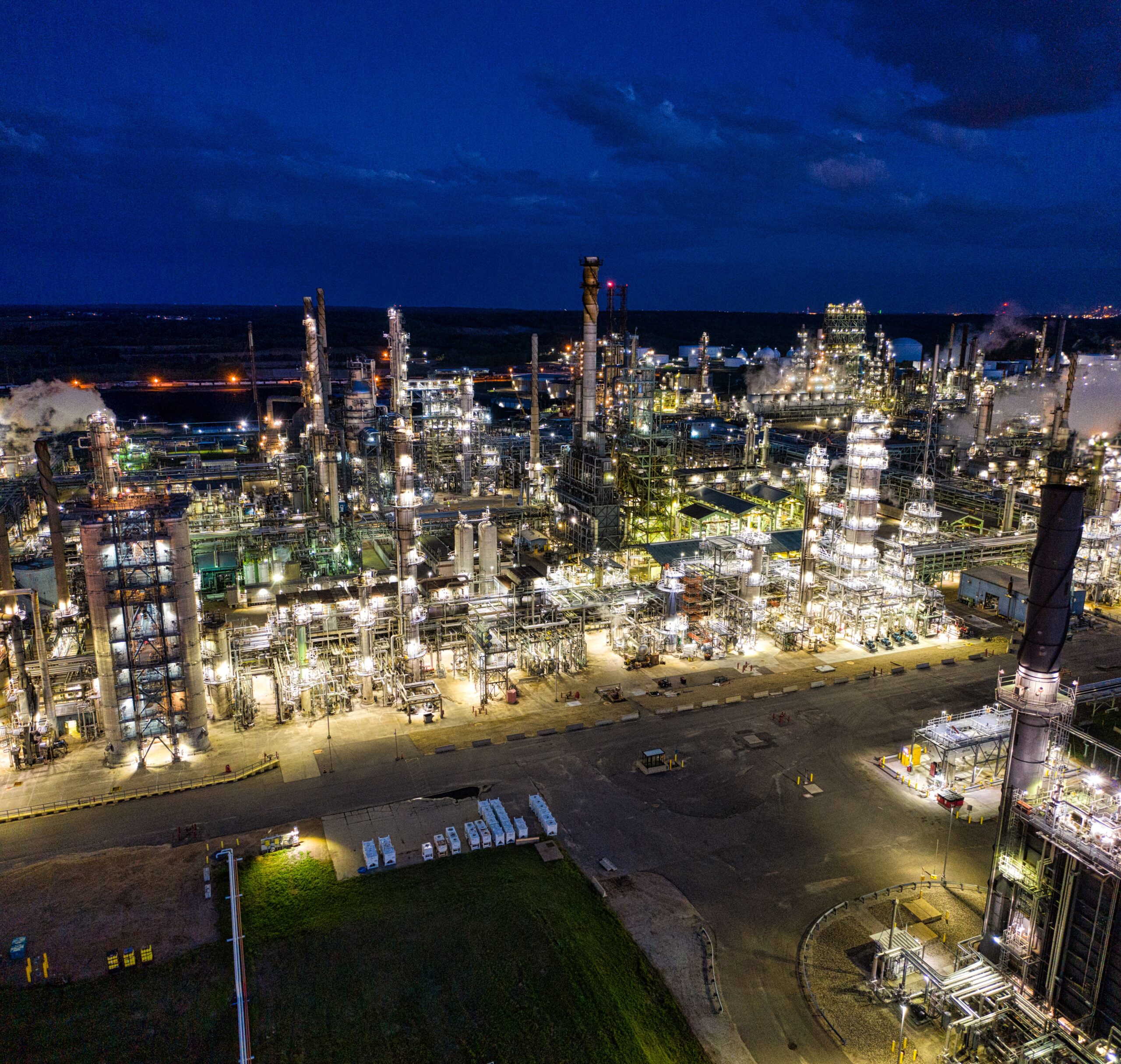 A professional financial journalism photograph showcasing massive industrial petrochemical energy processing pipeline networks illuminated under intense nocturnal security lighting