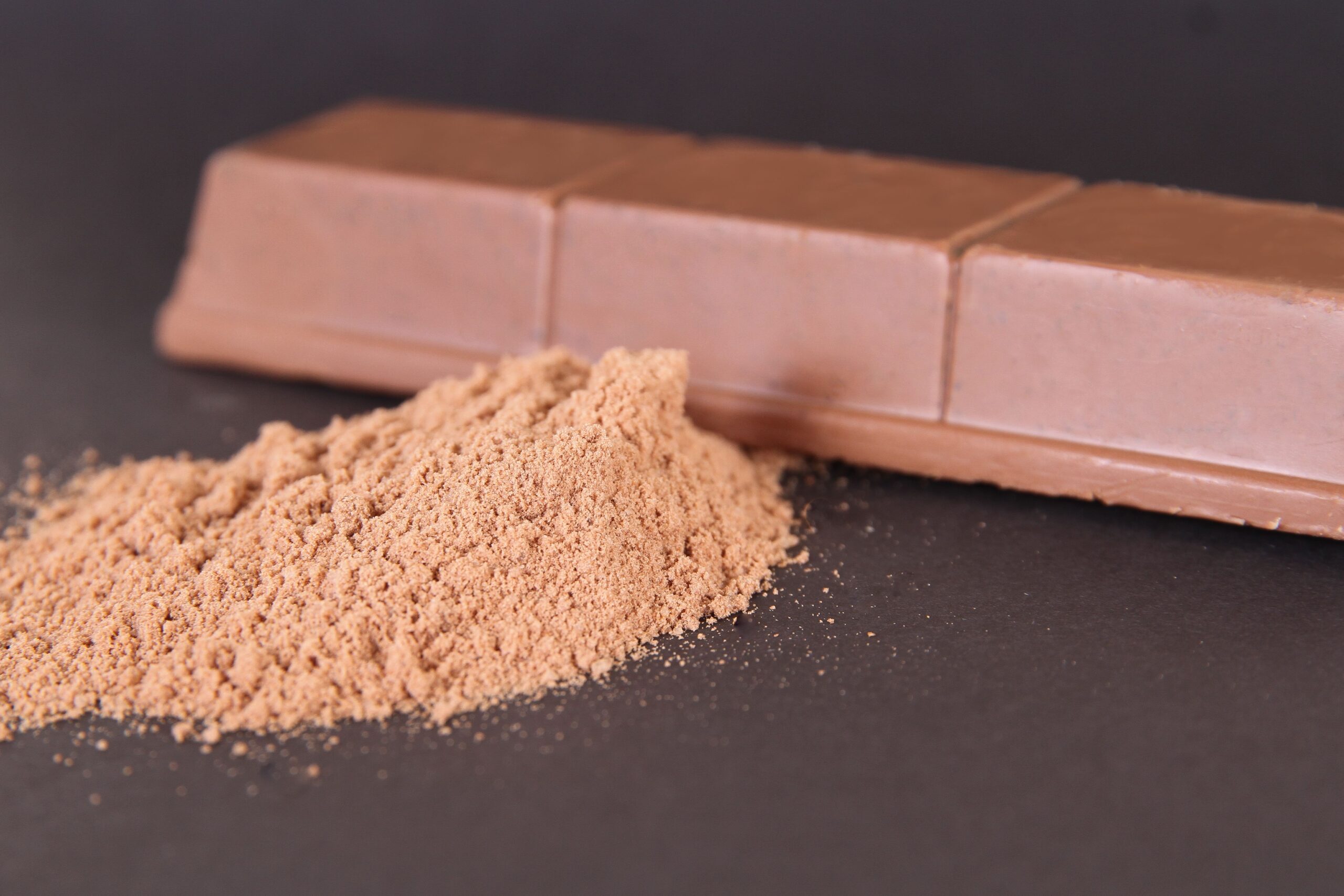 A professional commercial photograph displaying thick broken blocks of rich dark chocolate scattered next to raw cacao beans and powder