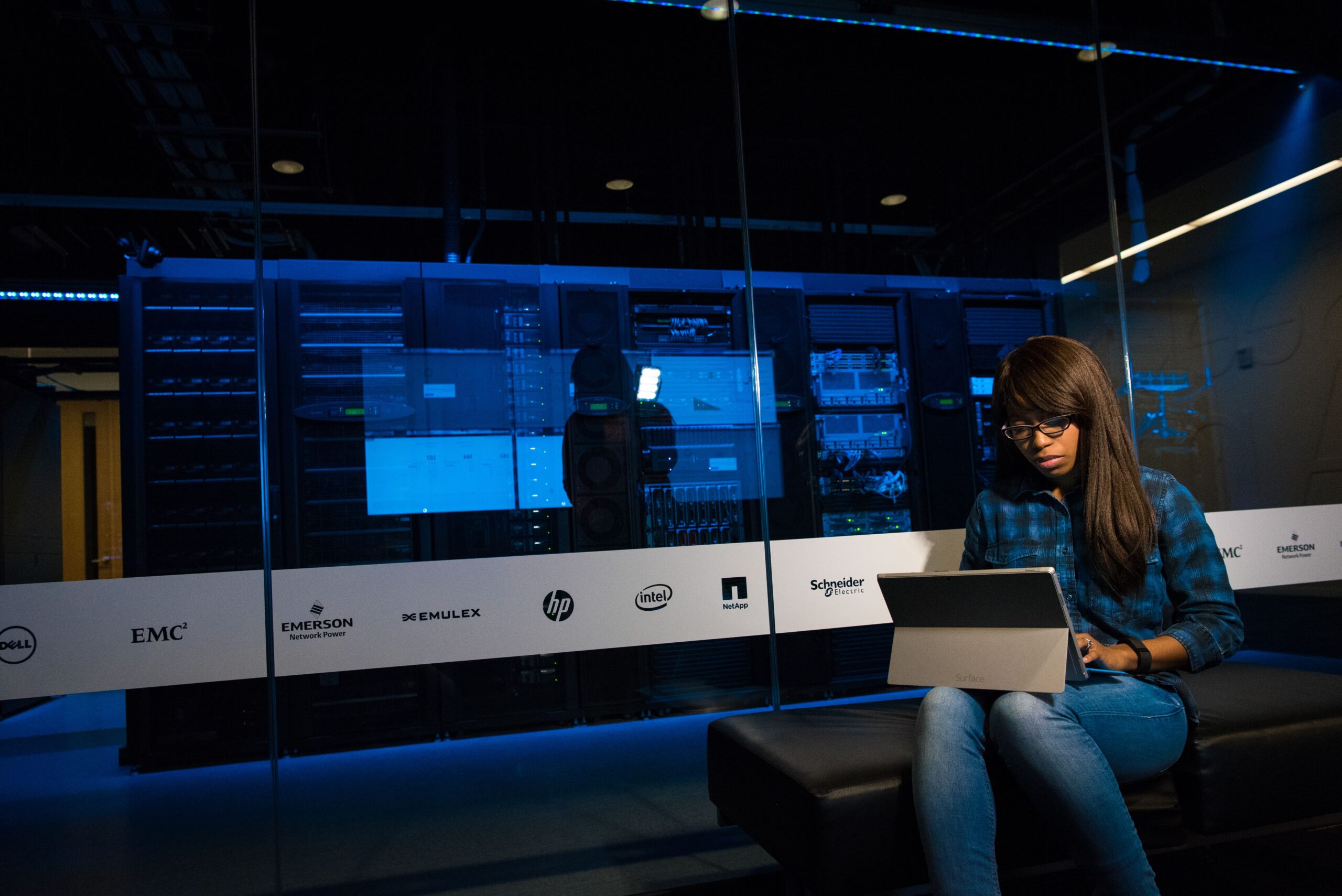 A modern industrial server rack room illuminated by dark neon blue LED status indicators with high-tech security overlay graphics